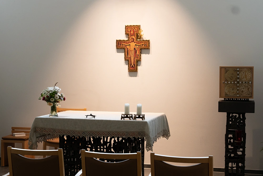Andachtsraum mit Kreuz und Altar Ein schlichter Andachtsraum mit einem Altar, auf dem zwei Kerzen und ein Blumenstrauß stehen. Dahinter hängt ein Kreuz mit Christusdarstellung an der Wand. Mehrere Stühle sind vor dem Altar aufgereiht.