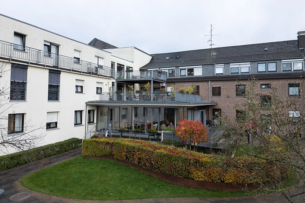 Blick auf das Regina Pacis mit heller und dunkler Fassade, Balkonen und einem gepflegten Gartenbereich mit Rasen und Hecken.