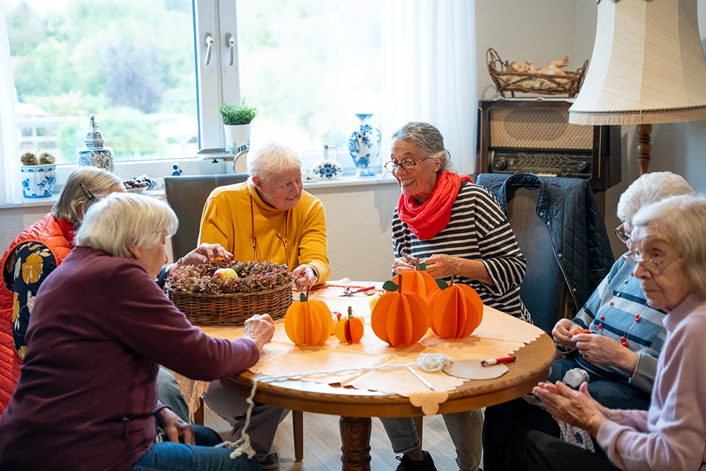 Eine Gruppe Senioren sitzt an einem runden Tisch und bastelt herbstliche Kürbisdekoration. Auf dem Tisch liegen Papierkürbisse, Scheren und ein Korb mit Trockenblumen. Der Raum ist gemütlich eingerichtet.