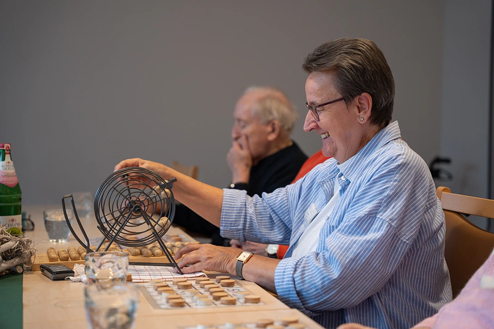 Betreuungskraft dreht Bingo-Trommel Eine Betreuungskraft mit Brille und gestreiftem Hemd sitzt an einem Tisch und dreht eine Bingo-Trommel. Auf dem Tisch liegen Spielchips, Wassergläser und Bingo-Karten. Im Hintergrund sitzen ältere Teilnehmer.