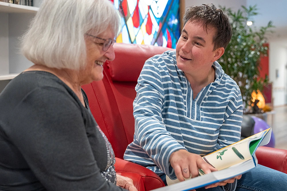 Lesestunde mit Seniorin im Franziskus-Haus Eine Betreuungsperson zeigt einer Seniorin ein geöffnetes Buch mit Illustrationen. Beide sitzen nebeneinander in roten Sesseln in einem Aufenthaltsbereich mit Bücherregal, Pflanzen und buntem Glasfenster.