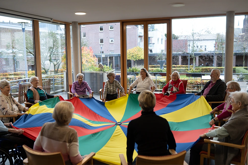Eine Gruppe Senioren sitzt in einem Kreis und hält ein buntes Schwungtuch in einem Raum mit großen Fenstern und Blick auf den Gartenbereich.