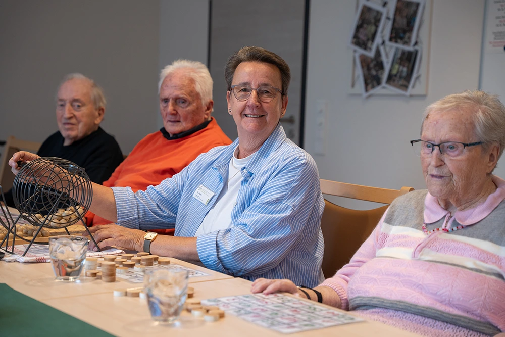 Bingo-Runde im Seniorenheim Drei Senioren und eine Betreuungsperson sitzen an einem Tisch im Innenraum und spielen Bingo. Die Betreuungsperson dreht die Bingo-Trommel, auf dem Tisch liegen Spielchips und Wassergläser.