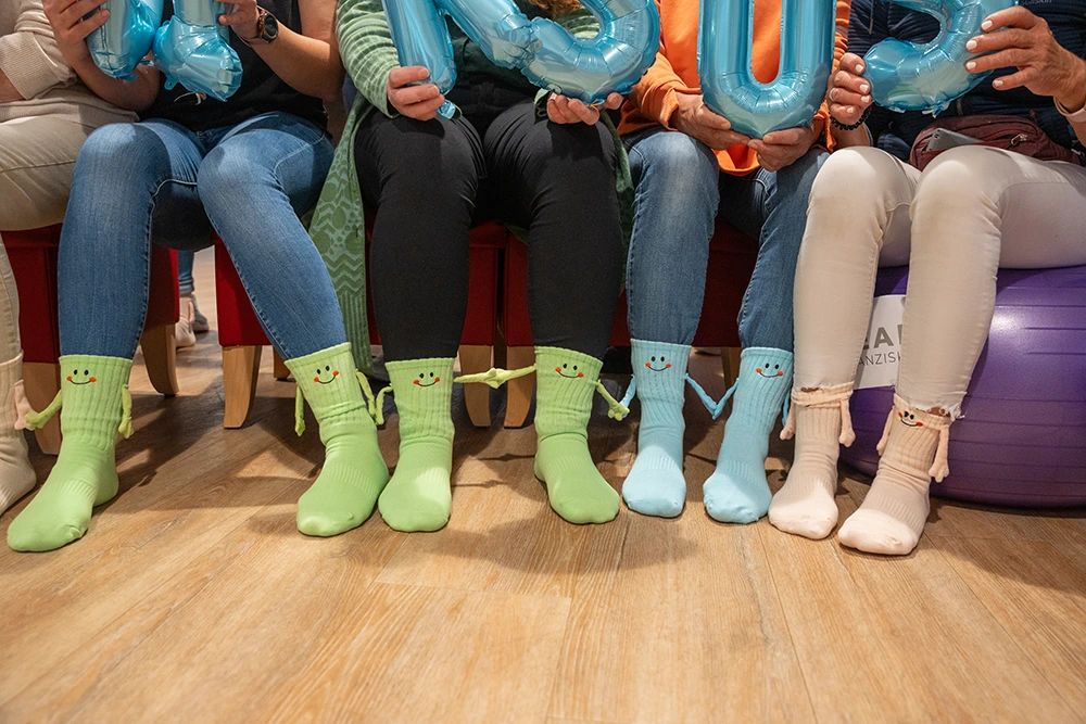 Aktionstag mit bunten Socken Mehrere Personen sitzen nebeneinander auf Stühlen und zeigen ihre bunten Socken mit lachenden Gesichtern und Armen. Über den Beinen halten sie blaue Luftballonbuchstaben.