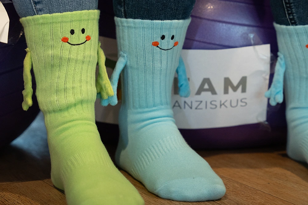 Nahaufnahme von zwei Paar bunten Socken mit aufgestickten lachenden Gesichtern und kleinen Armen. Im Hintergrund ist ein Gymnastikball mit der Aufschrift „Team Franziskus“ zu sehen.
