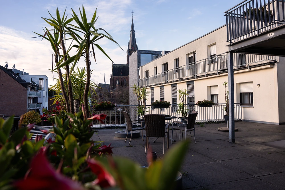 Eine begrünte Dachterrasse mit Tischen, Stühlen und Geländer, im Hintergrund ein Kirchturm und benachbarte Gebäude.