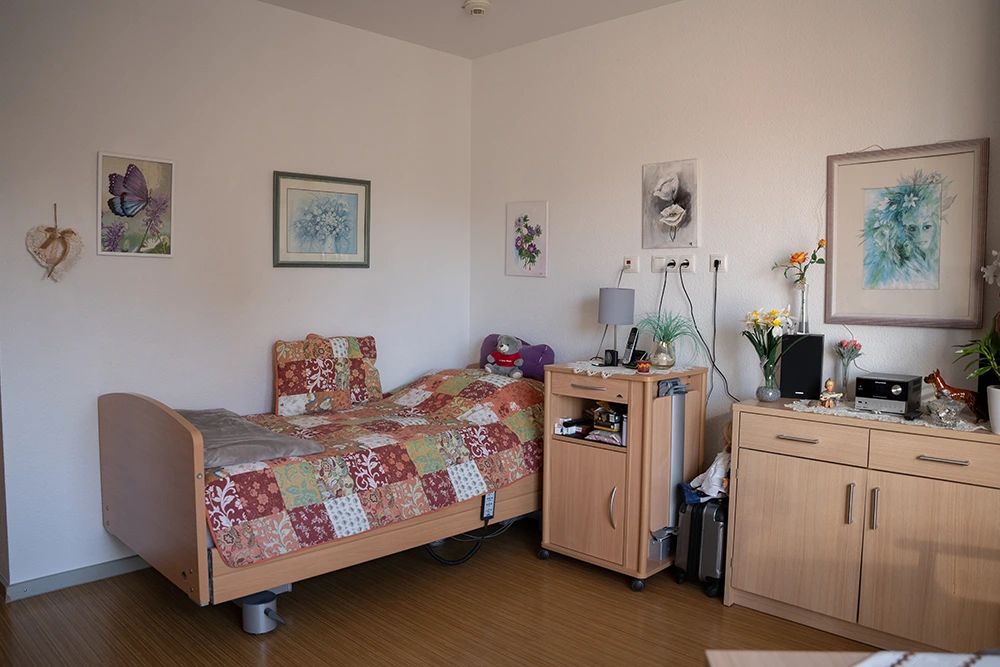 Ein möbliertes Einzelzimmer mit Pflegebett, Nachttisch, Kommode, Wandbildern und dekorativen Elementen an den Wänden.