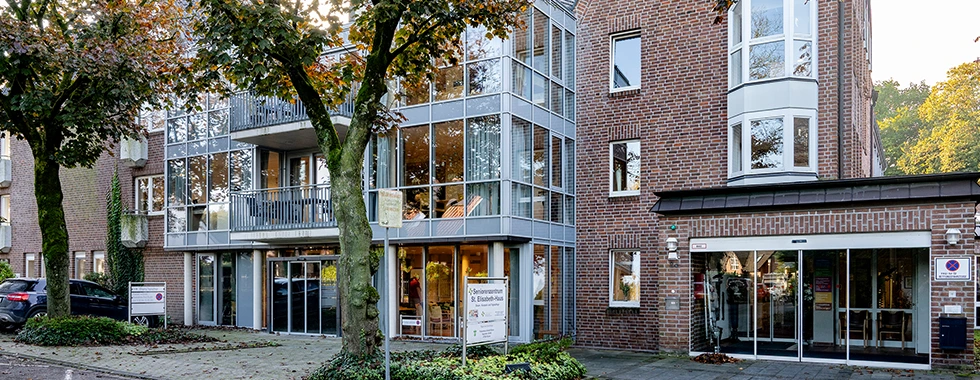 Mehrstöckiges Seniorenzentrum „St. Elisabeth-Haus“ mit großer Glasfassade, roten Backsteinelementen, Bäumen mit Herbstlaub im Vordergrund sowie ausgeschildertem Eingangsbereich.
