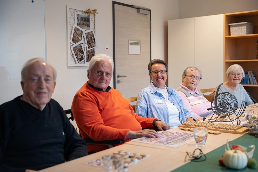 Fünf ältere Menschen sitzen an einem Tisch mit Bingo-Karten und einem Bingo-Gerät. Sie blicken freundlich in die Kamera, im Raum sind Dekoration und Möbel zu sehen.