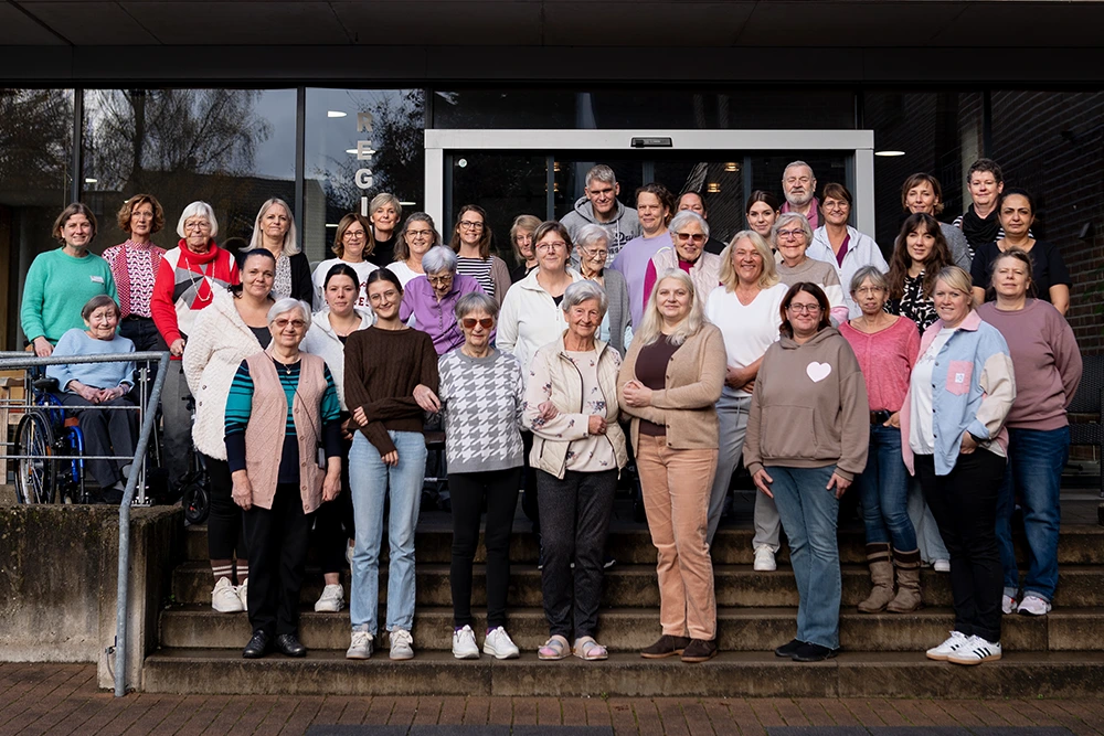 Eine große Gruppe von Mitarbeitenden und Senioren steht vor dem Eingangsbereich des Regina Pacis und blickt in die Kamera; einige Personen sitzen, andere stehen auf der Treppe.