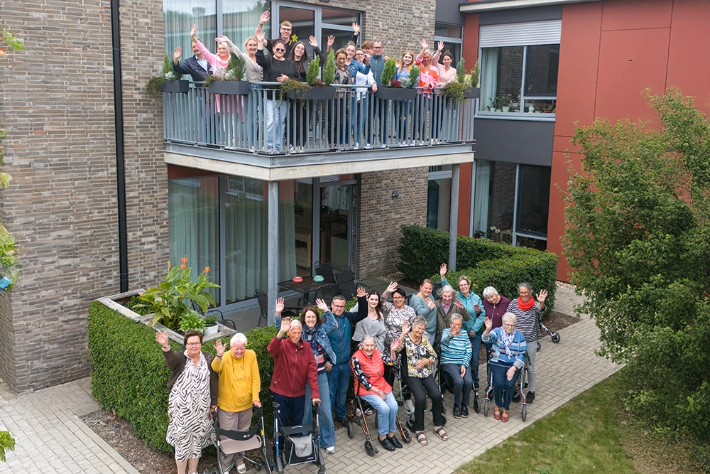 Eine Gruppe von Senioren und Betreuungspersonen steht und sitzt im Innenhof eines modernen Seniorenheims. Auf dem Balkon darüber winkt ein weiteres Team in die Kamera.