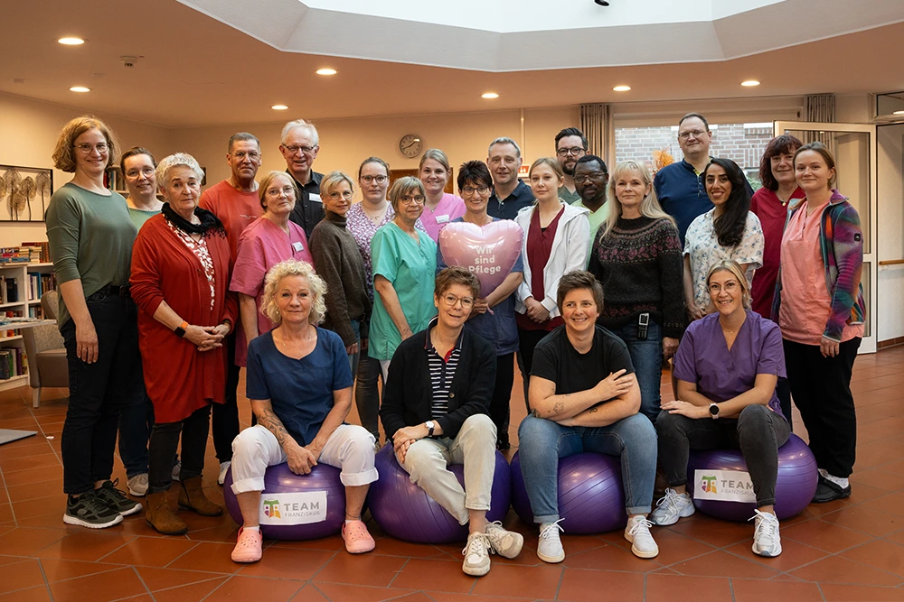 Eine große Gruppe von Pflegekräften und Mitarbeitenden steht und sitzt gemeinsam im Innenraum, einige sitzen auf Gymnastikbällen mit Team-Logo, in der Mitte wird ein Herzballon mit der Aufschrift „Wir sind Pflege“ gehalten.