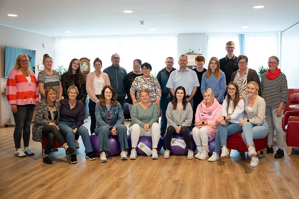 Gruppenfoto Team Veronika Innenraum Eine große Gruppe von Mitarbeitenden sitzt und steht in einem Aufenthaltsraum. Einige Personen sitzen auf Gymnastikbällen, im Hintergrund befinden sich Pflanzen, eine Uhr und große Fenster.