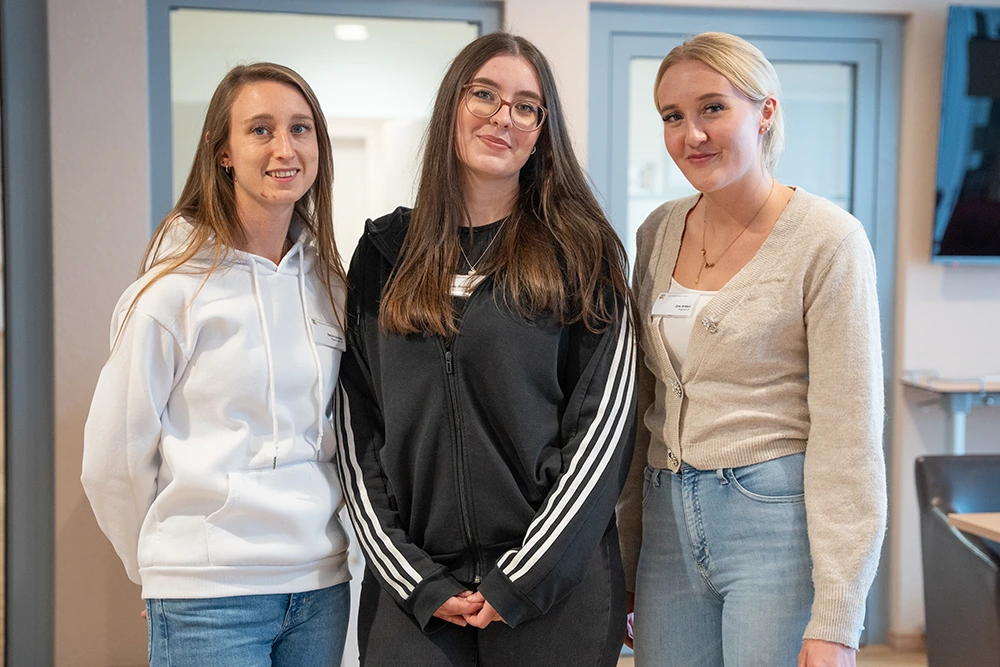 Drei junge Mitarbeiterinnen im Portrait Drei junge Frauen stehen nebeneinander im Innenbereich eines Gebäudes und lächeln in die Kamera. Sie tragen Freizeitkleidung und Namensschilder. Im Hintergrund befinden sich Türen und Fenster.