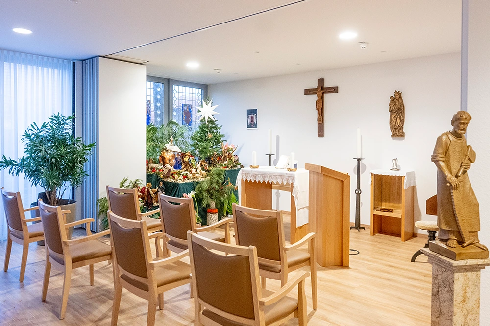 Kapelle mit Weihnachtskrippe Ein kleiner Kapellenraum mit Stühlen, Altar, Krippe und Weihnachtsdekoration. An der Wand hängen ein Kreuz und religiöse Figuren, der Raum ist hell und freundlich gestaltet.