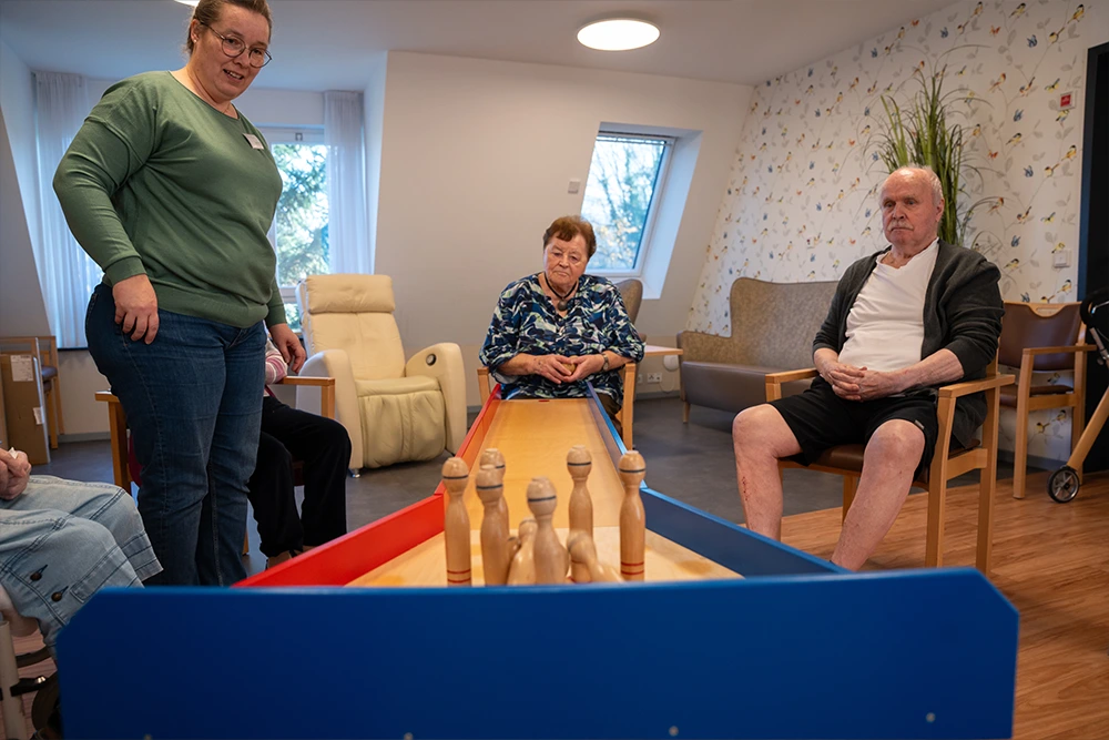 Eine Pflegekraft betreut mehrere Seniorinnen und Senioren beim Spielen mit einem Kegelspiel an einem Tisch. Alle sitzen im Kreis in einem wohnlich eingerichteten Raum.