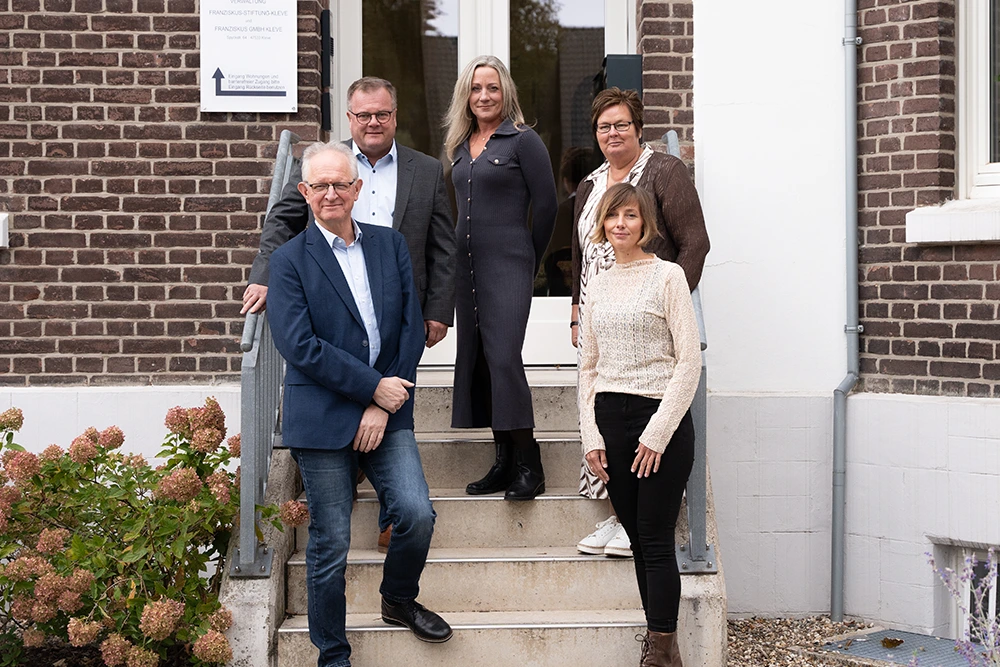 Fünf Personen stehen auf einer Außentreppe vor einem Gebäude mit Backsteinfassade und weißem Türrahmen. Links ist ein Hinweisschild mit dem Namen „Franziskus GmbH Kleve“ zu sehen. Zwei Männer und drei Frauen sind frontal zur Kamera ausgerichtet und lächeln.