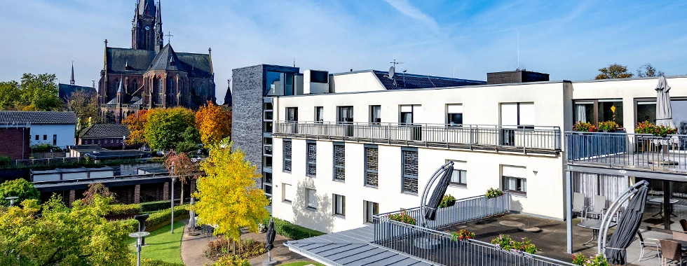 Helles, mehrstöckiges Seniorenwohnhaus „Regina Pacis“ in Kevelaer mit mehreren Balkonen und bepflanzten Terrassen, im Hintergrund eine große Kirche mit Türmen und herbstlich gefärbten Bäumen.