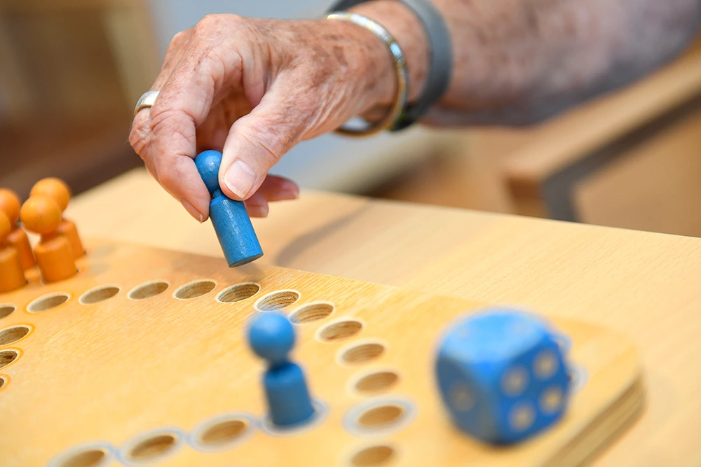 Senior spielt Brettspiel mit Figuren Eine Hand setzt eine blaue Spielfigur auf ein Holzbrettspiel mit eingefrästen Spielfeldern. Auf dem Tisch liegen weitere Spielfiguren und ein großer blauer Holzwürfel.