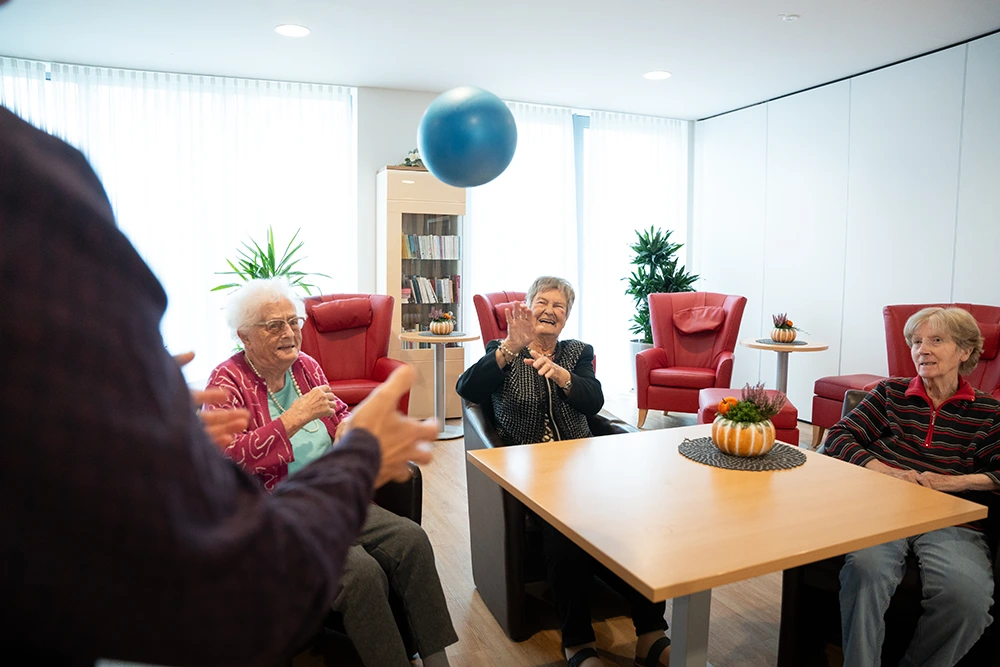 Drei Seniorinnen sitzen in Sesseln und werfen sich einen blauen Ball zu. Eine weitere Person steht im Vordergrund. Der Raum ist hell mit roten Sesseln, Pflanzen und herbstlicher Dekoration.