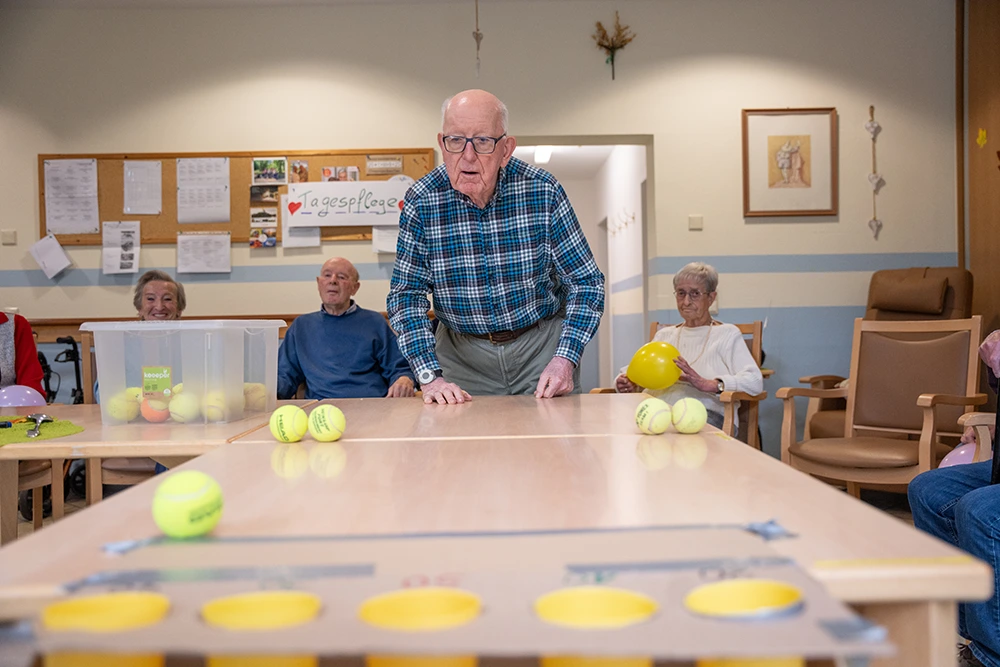 Ein Senior steht an einem langen Tisch und spielt ein Bewegungsspiel mit mehreren Tennisbällen. Um ihn herum sitzen weitere Senioren mit bunten Luftballons in einem Aktivitätsraum.