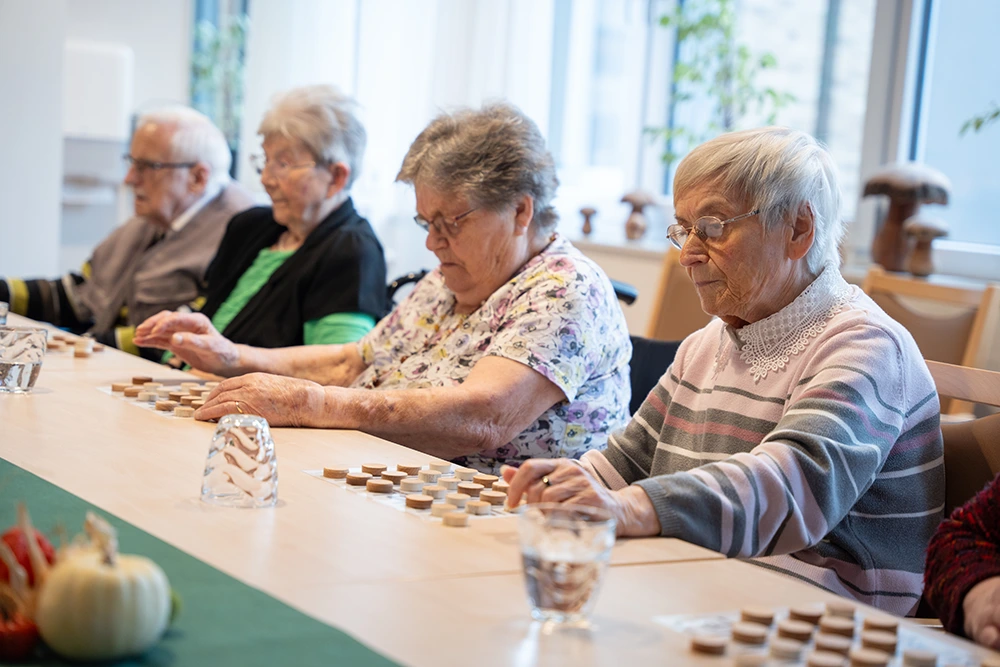 Senioren spielen Memory am Tisch Vier ältere Menschen sitzen an einem Tisch und spielen ein Memory-Spiel mit runden Holzplättchen. Auf dem Tisch stehen mehrere Gläser mit Wasser. Die Szene spielt sich in einem hellen Raum mit großen Fenstern ab.