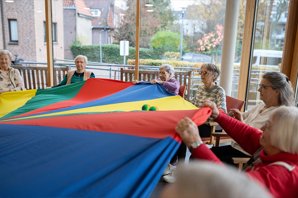 Eine Gruppe Senioren sitzt im Kreis und hält gemeinsam ein großes, buntes Schwungtuch in einem Raum mit großen Fenstern.
