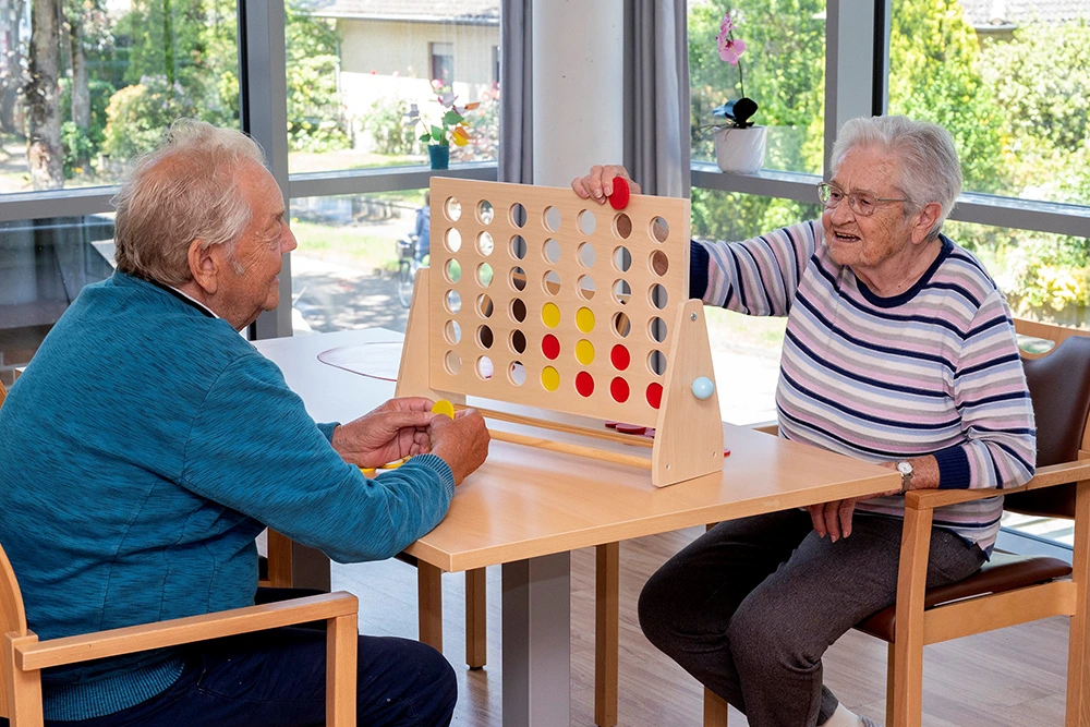 Senioren spielen Vier gewinnt Ein Senior und eine Seniorin sitzen sich an einem Tisch gegenüber und spielen eine Partie Vier gewinnt mit roten und gelben Spielsteinen. Sie befinden sich in einem Raum mit großen Fenstern und Blick ins Grüne.