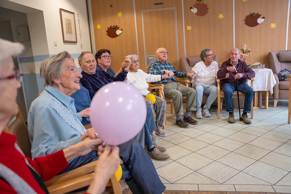 Eine Gruppe Senioren sitzt in einem Stuhlkreis und hält sich an den Händen, während eine Person einen rosafarbenen Luftballon in der Hand hält. Der Raum ist herbstlich dekoriert mit Wandmotiven.