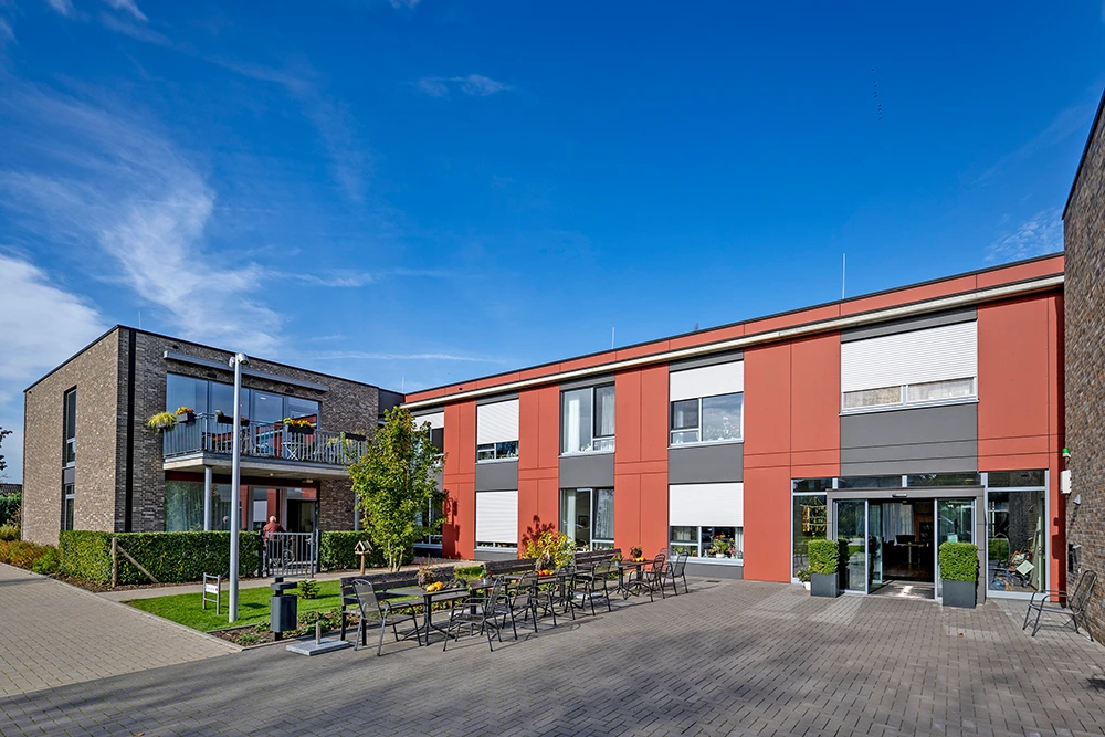 Moderner Gebäudekomplex mit roter und grauer Fassade bei sonnigem Wetter. Im Innenhof stehen mehrere Tische und Stühle, einige Balkone sind bepflanzt. Der Himmel ist blau mit wenigen Wolken.