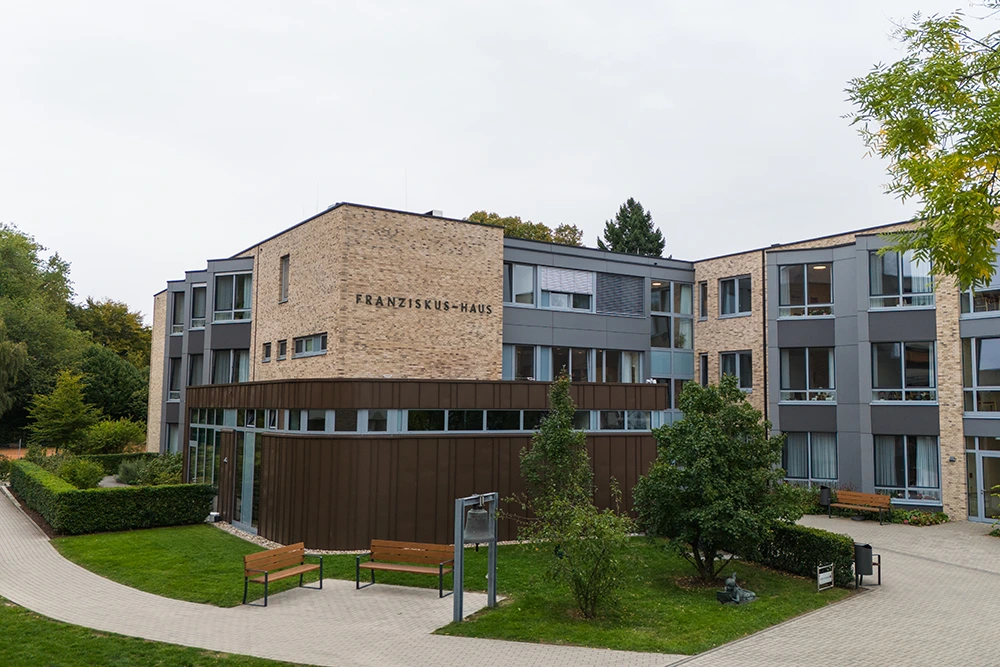 Modernes Seniorenheim mit der Aufschrift „Franziskus-Haus“ auf der Fassade, umgeben von Wegen, Bäumen, Bänken und gepflegtem Grün. Das Gebäude ist mehrstöckig und besteht aus einer Kombination von Ziegel- und Metallfassaden.