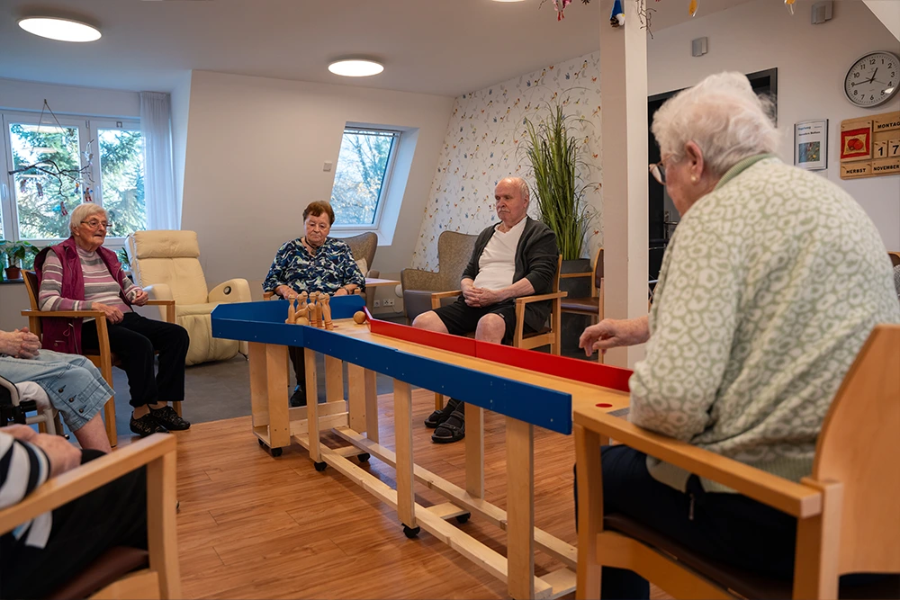 Mehrere Senioren sitzen im Kreis in einem Aufenthaltsraum und spielen mit einem Tisch-Kegelspiel. Der Raum ist freundlich eingerichtet mit Sesseln, Pflanzen und großen Fenstern.