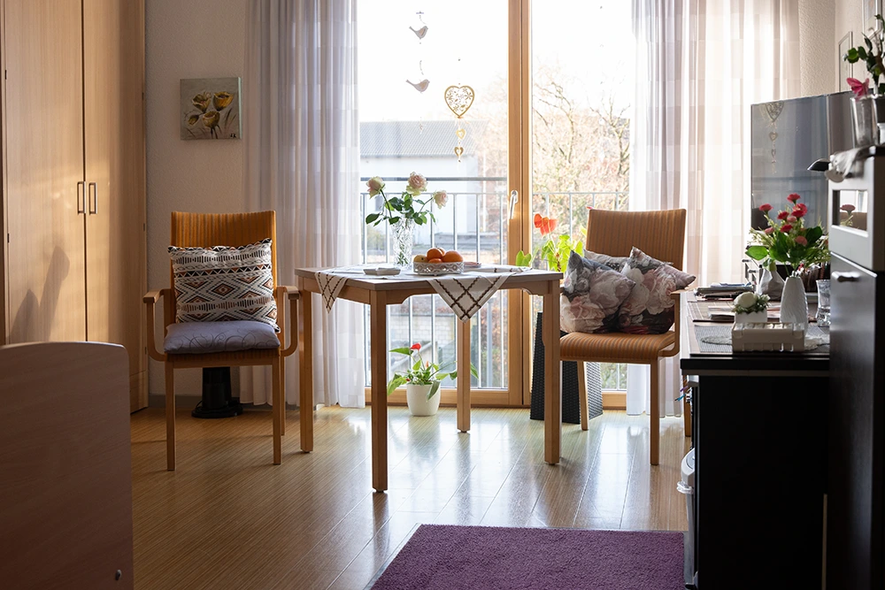 Ein heller Wohnraum mit zwei Holzstühlen, einem kleinen Tisch mit Tischdecke und Pflanzen, großen Fenstern mit Vorhängen sowie einem Schrank und einem Fernseher.