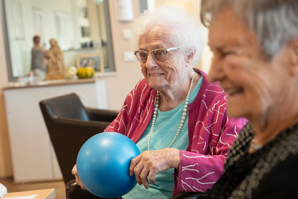 Seniorin mit Ball beim Spiel Eine Seniorin mit weißem Haar, Brille und pinker Strickjacke sitzt auf einem Sessel und hält einen blauen Ball in den Händen. Neben ihr sitzt eine weitere lächelnde Frau. Im Hintergrund ist ein Spiegel sichtbar.