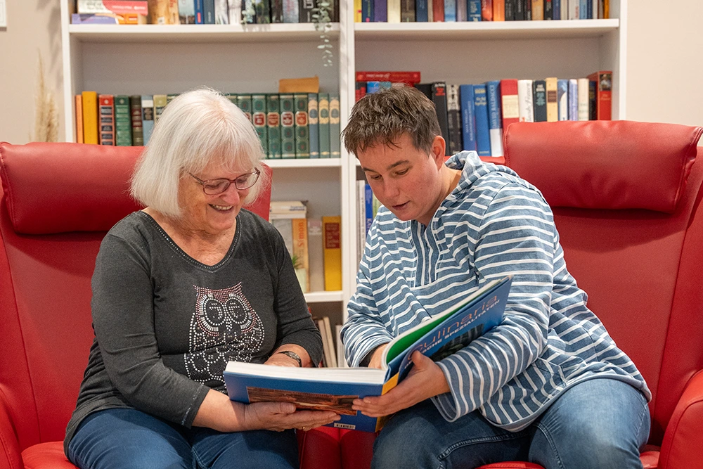 Eine ältere Frau und eine jüngere Person sitzen auf roten Sesseln und schauen gemeinsam in ein aufgeschlagenes Buch. Im Hintergrund steht ein Bücherregal.