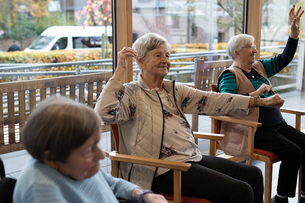 Drei ältere Frauen machen sitzend Armübungen in einem hellen Raum mit Fensterfront; eine der Frauen trägt eine beige Weste und lächelt.