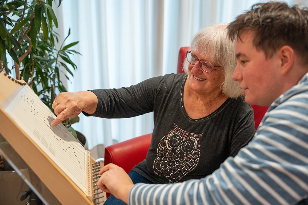 Eine Seniorin sitzt mit einer Betreuungsperson vor einem Klanginstrument mit Notenvorlage und zeigt auf die Saiten. Beide lächeln, im Hintergrund stehen ein roter Sessel und eine Pflanze.