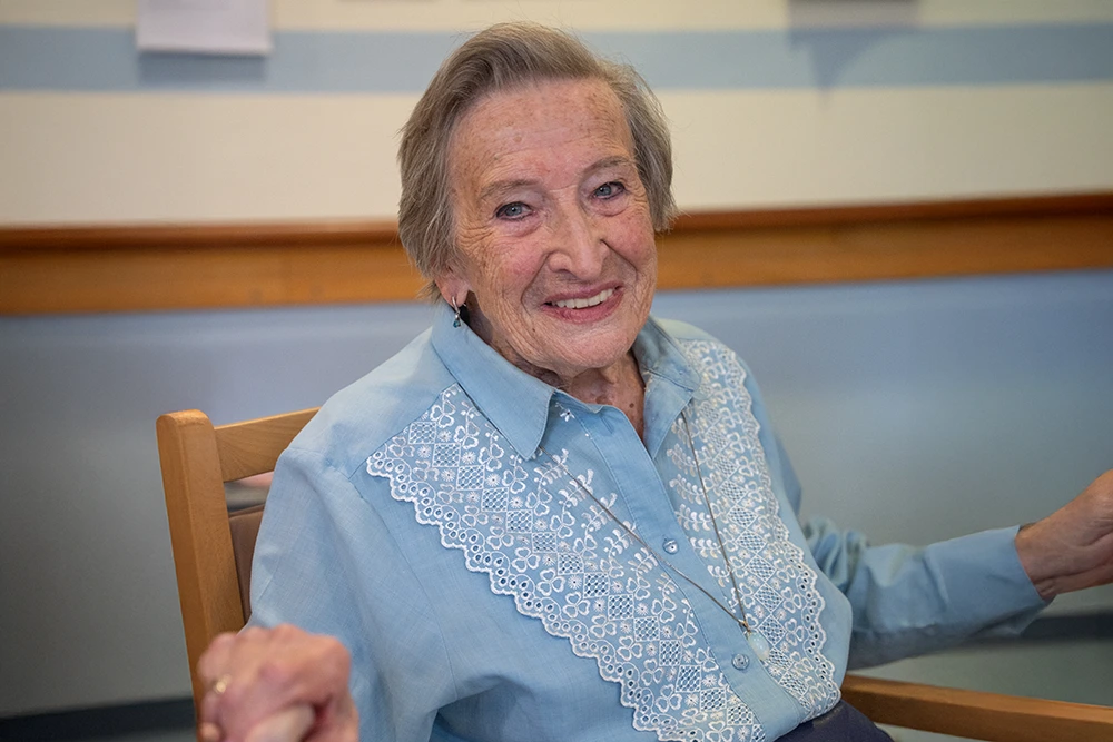 Lächelnde Seniorin im Sessel Eine Seniorin mit grauen Haaren und hellblauer Bluse mit weißer Stickerei sitzt auf einem Holzstuhl und lächelt in die Kamera. Der Hintergrund zeigt eine Wand mit Streifenmuster.