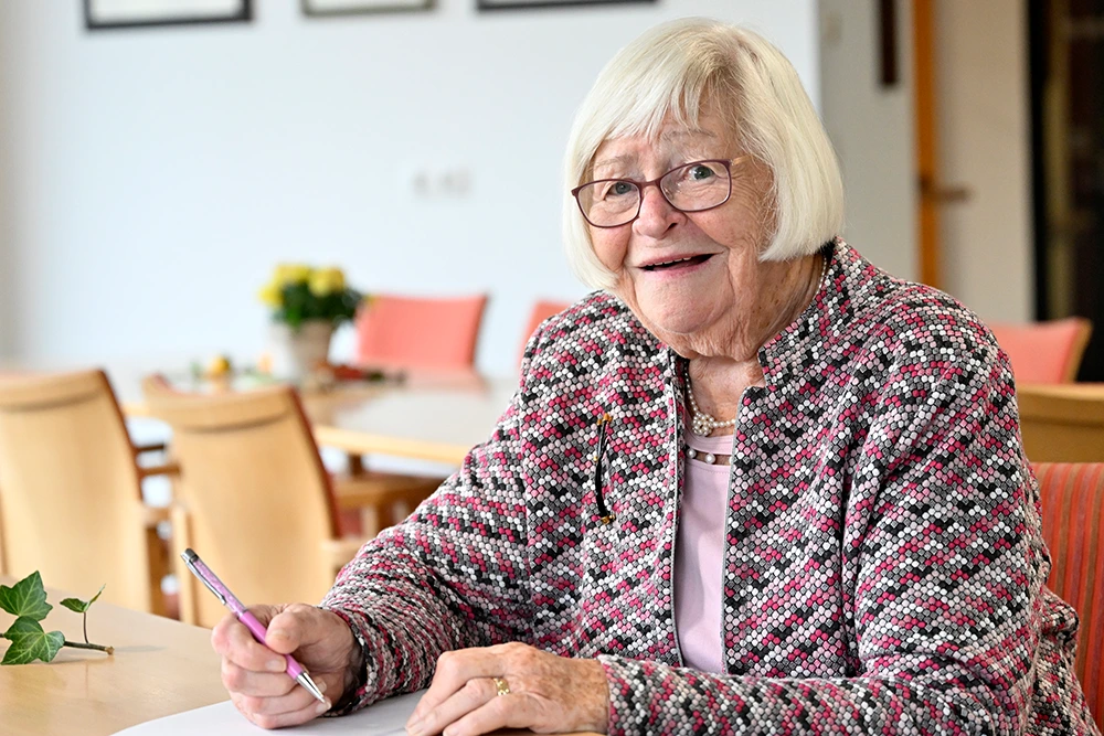 Seniorin beim Schreiben am Tisch Eine Seniorin mit weißem Haar, Brille und gemusterter Jacke sitzt lächelnd an einem Tisch mit Stift und Papier in der Hand. Im Hintergrund stehen Stühle und Tische in einem Gemeinschaftsraum.