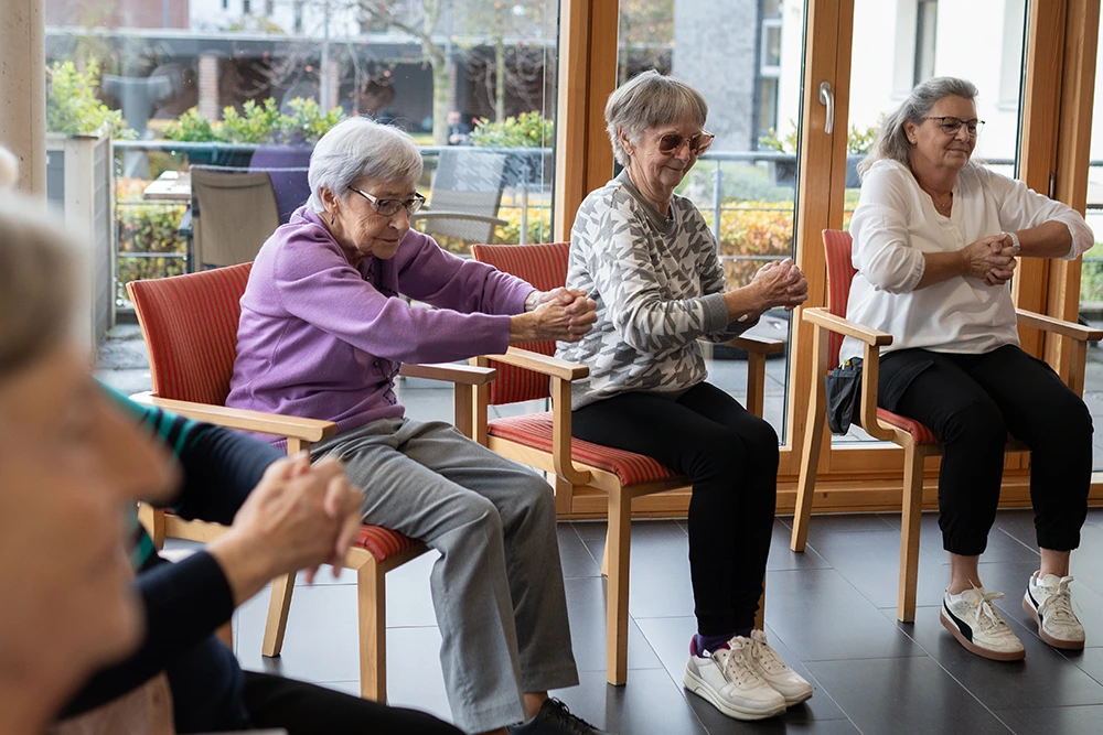 Drei ältere Frauen sitzen auf Stühlen und führen synchron eine Armübung mit gefalteten Händen durch, in einem hellen Raum mit bodentiefen Fenstern.