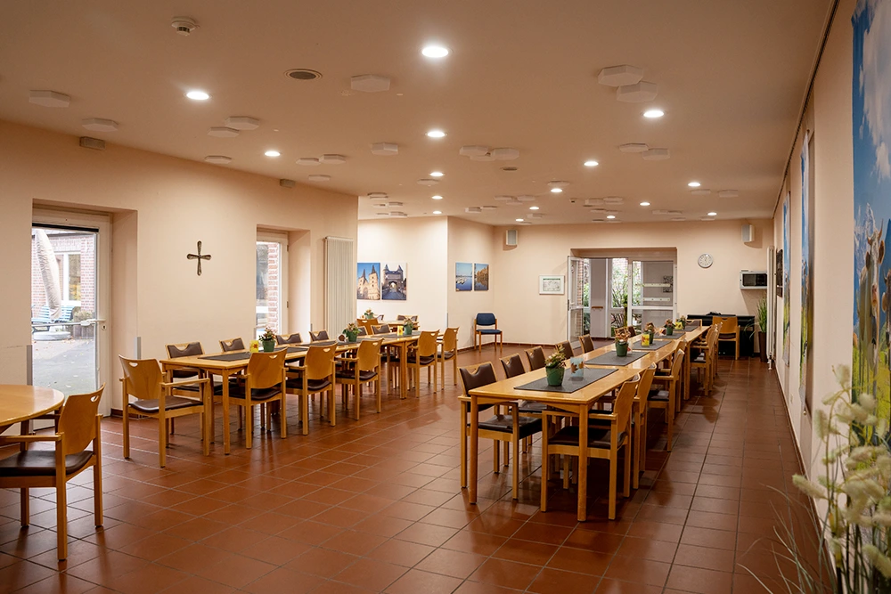 Langer, gut beleuchteter Speisesaal mit vielen gedeckten Holztischen und Stühlen, Pflanzen- und Landschaftsbildern an den Wänden sowie Zugang nach draußen.