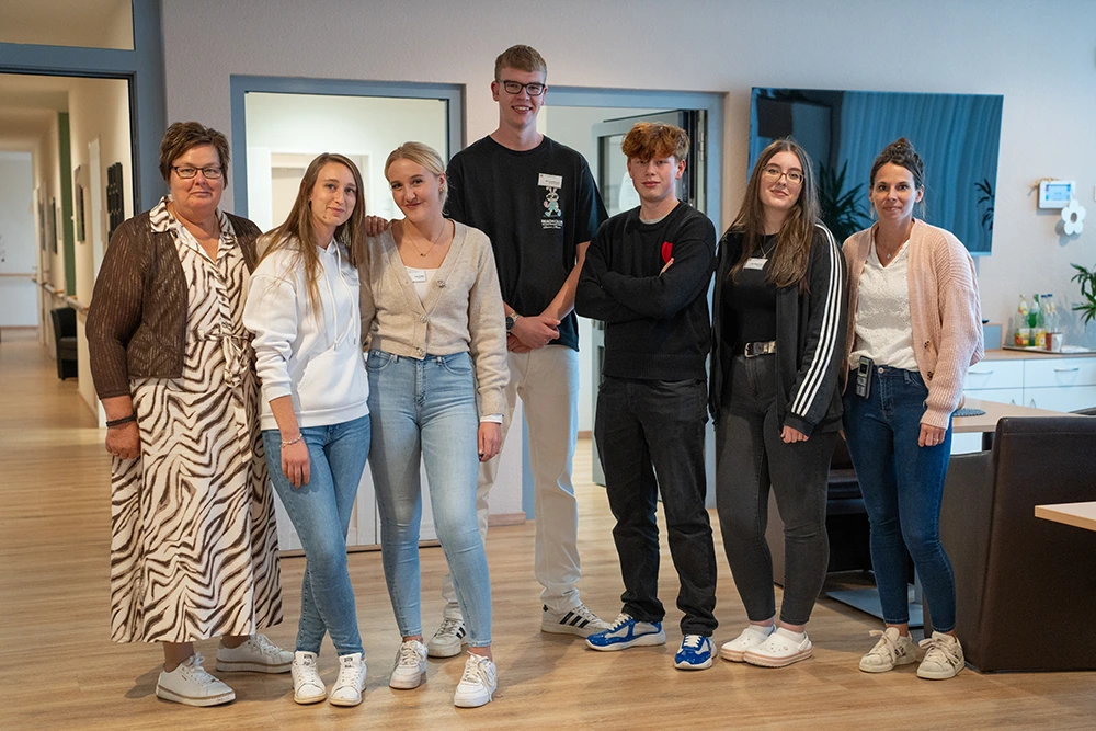 Sieben Personen unterschiedlichen Alters stehen in einem Flur und posieren für ein Gruppenfoto. Im Hintergrund sind Türen und ein heller Gemeinschaftsraum sichtbar.