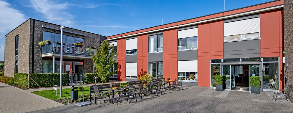 Das moderne „Veronika-Haus“ in Kleve-Rindern mit zweistöckigem Gebäude, roter und grauer Fassade, großen Fenstern und einem gepflasterten Hof mit zahlreichen Stühlen und Tischen im Freien.