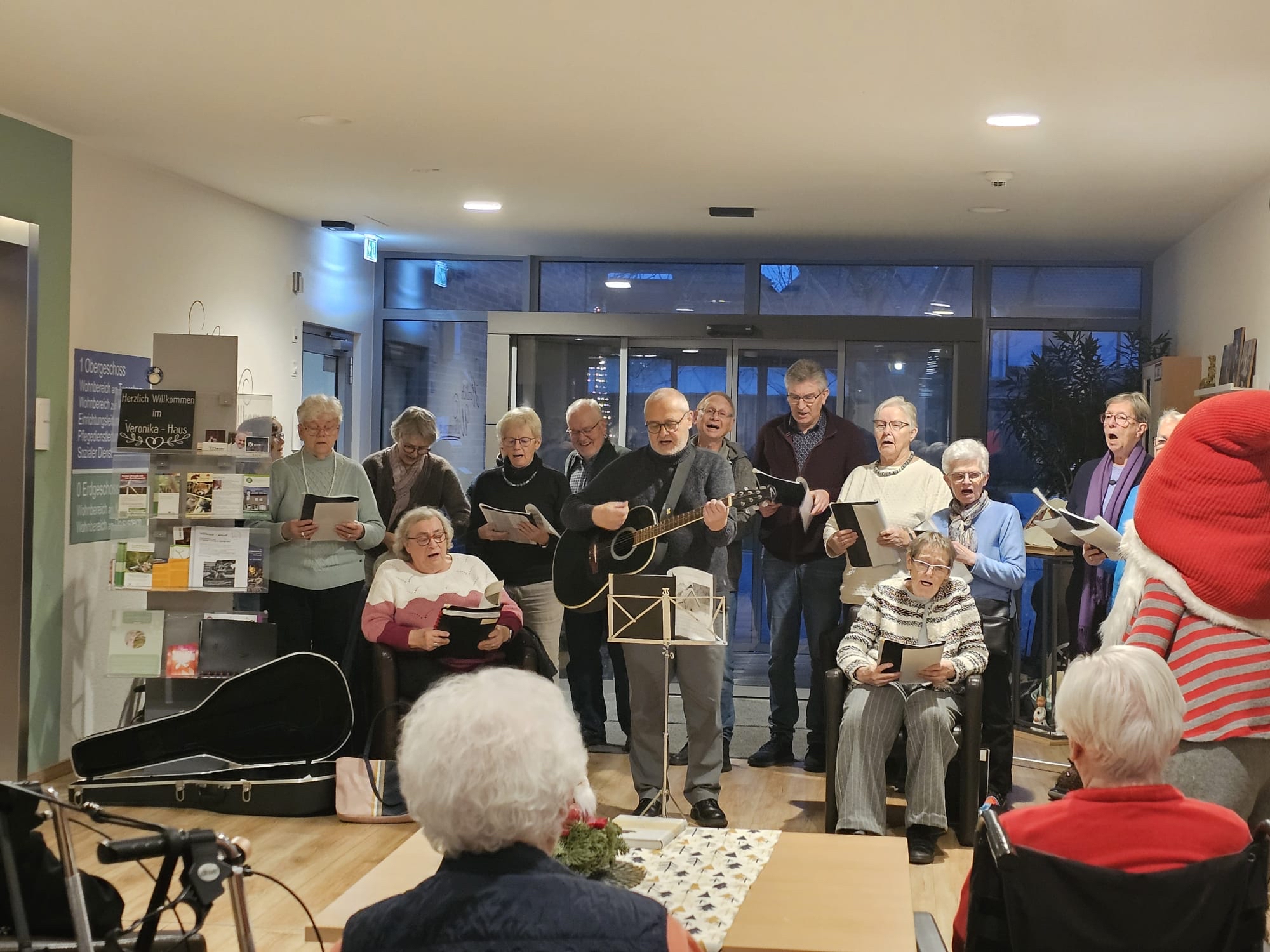 Eine Gruppe älterer Menschen singt gemeinsam in einem Seniorenheim. Einige stehen, andere sitzen im Rollstuhl, eine Person spielt Gitarre, Notenblätter werden gehalten.