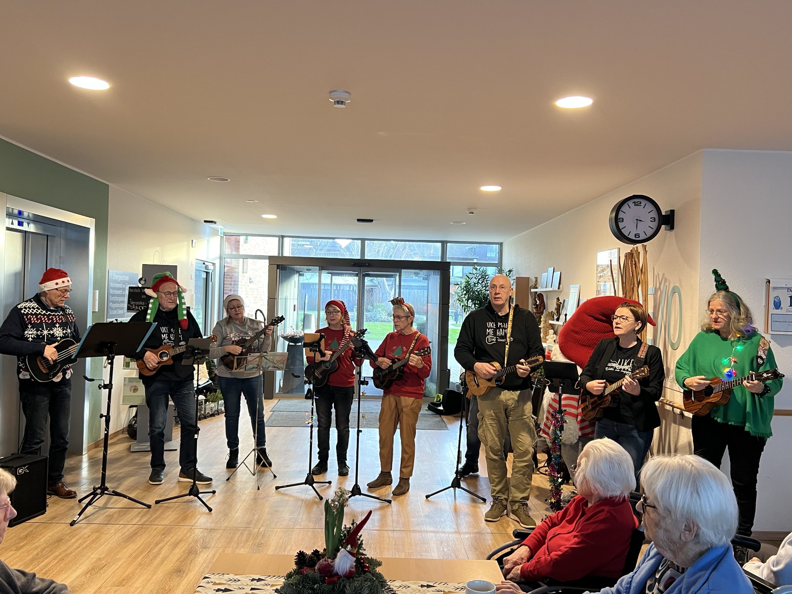 Eine Gruppe von Erwachsenen mit Ukulelen, teils in Weihnachtskleidung, spielt Musik in einer Seniorenresidenz. Senioren sitzen davor und hören zu.