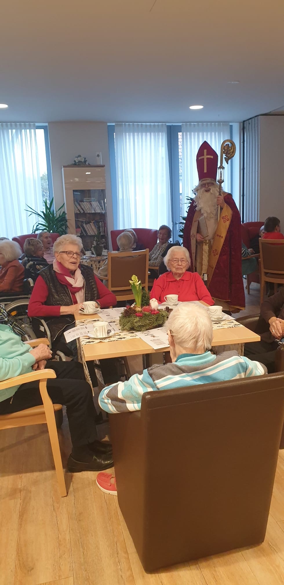 Senioren sitzen an gedeckten Tischen im Gemeinschaftsraum eines Heims, im Hintergrund steht eine Person im Nikolauskostüm mit Bischofsstab.