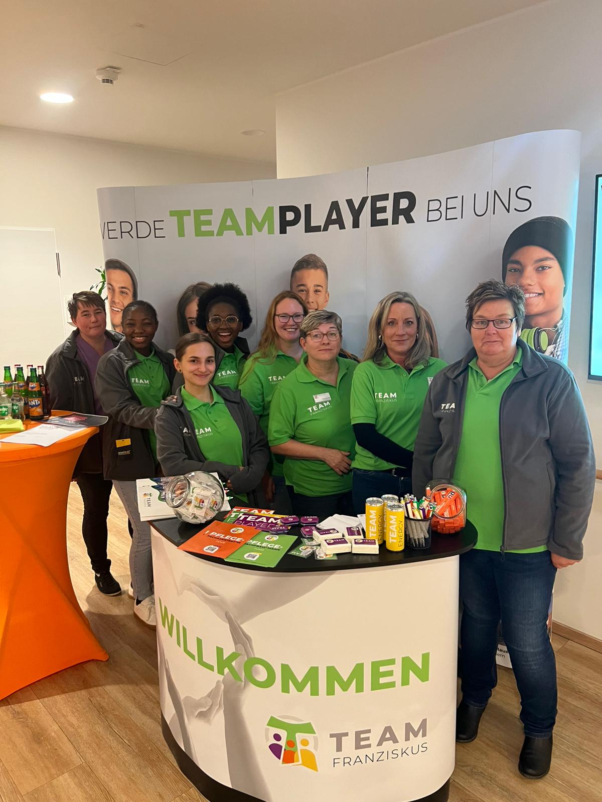 Team Franziskus am Infostand Mehrere Mitarbeiterinnen von Team Franziskus stehen an einem Infostand mit Werbematerialien und einem „Willkommen“-Schriftzug. Alle tragen grüne Polohemden mit dem Team-Logo. Im Hintergrund befindet sich ein großes Banner mit der Aufschrift „Werde Teamplayer bei uns“ und Porträts verschiedener Personen. Links steht ein Stehtisch mit Getränken.
