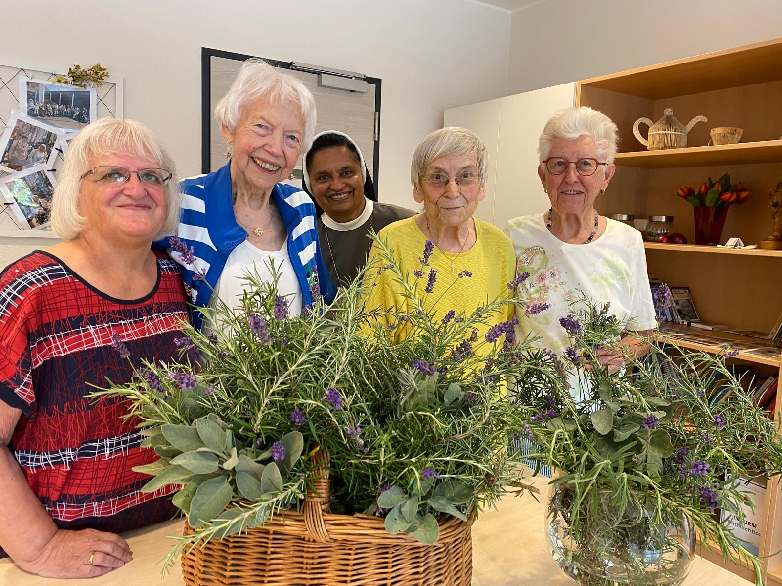 Fünf ältere Frauen, darunter eine Ordensschwester, stehen gemeinsam hinter einem Tisch mit zwei großen Gefäßen voller frischer Kräutersträuße, darunter Lavendel, Rosmarin und Salbei. Sie lächeln in die Kamera, im Hintergrund sind ein Regal mit Dekoration und eine Pinnwand mit Fotos zu sehen.