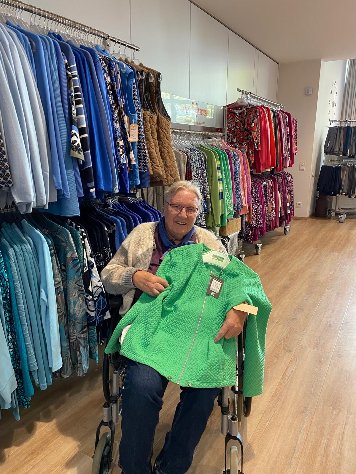 Eine ältere Frau im Rollstuhl hält eine grüne Jacke mit Preisschild auf dem Schoß und sitzt in einem Bekleidungsgeschäft. Um sie herum hängen bunte Kleidungsstücke, darunter Jacken, Pullover und Blusen in verschiedenen Farben. Der Boden ist aus hellem Holz, die Umgebung ist hell und ordentlich.