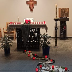 Ein festlich geschmückter Altar in einer Kapelle mit vielen roten Kerzen, dunklen Tüchern, Pflanzen und einem Herz aus Stoff und Kerzen auf dem Boden. Im Hintergrund ein Kreuz an der Wand und ein Tabernakel.